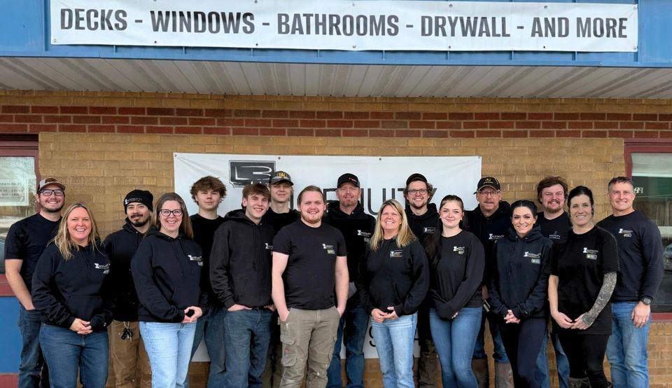 A group of 17 workers in matching black shirts pose in front of a building with a construction service sign.