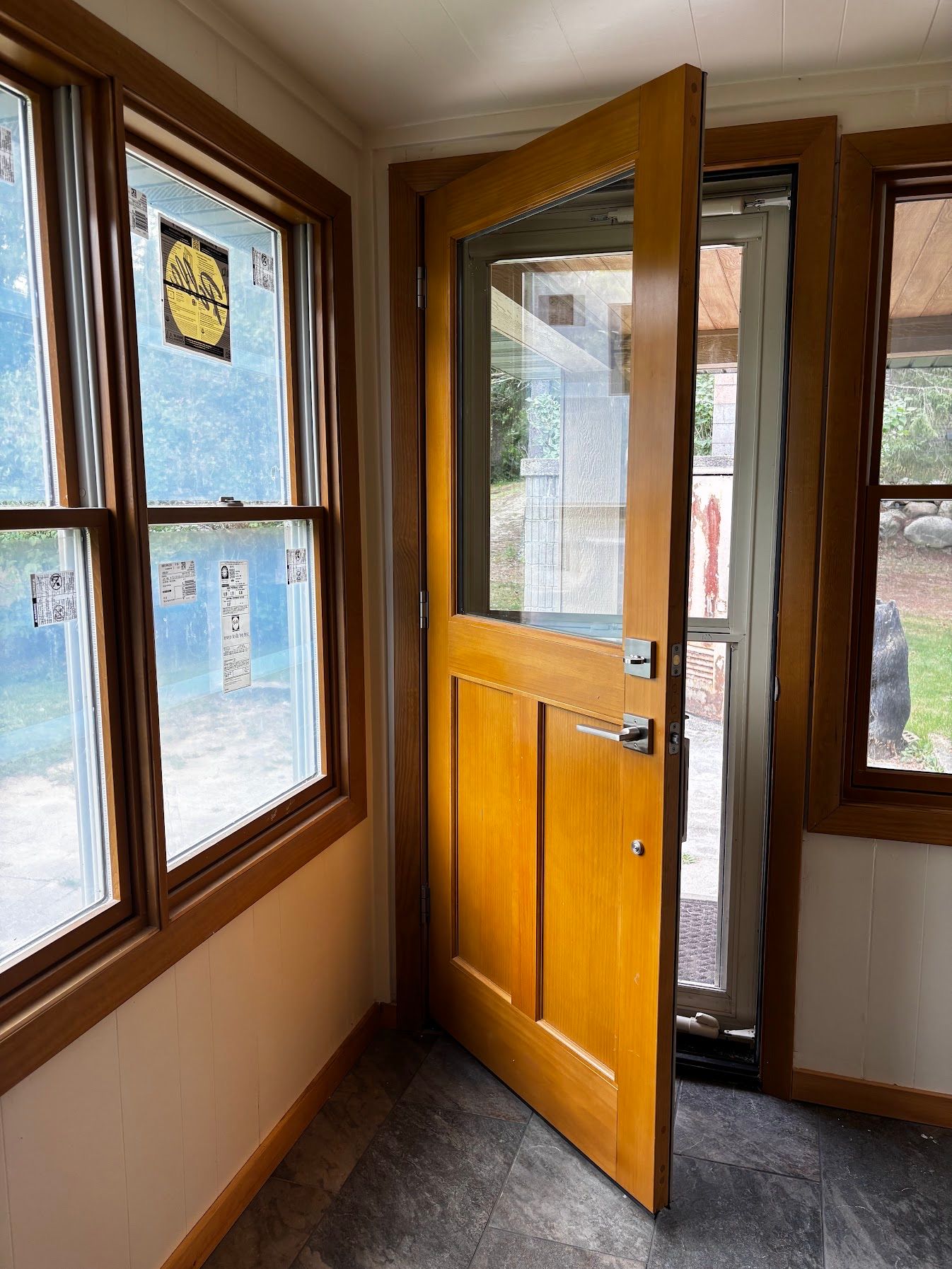 Wooden front door ajar in a small entryway with windows and daylight outside