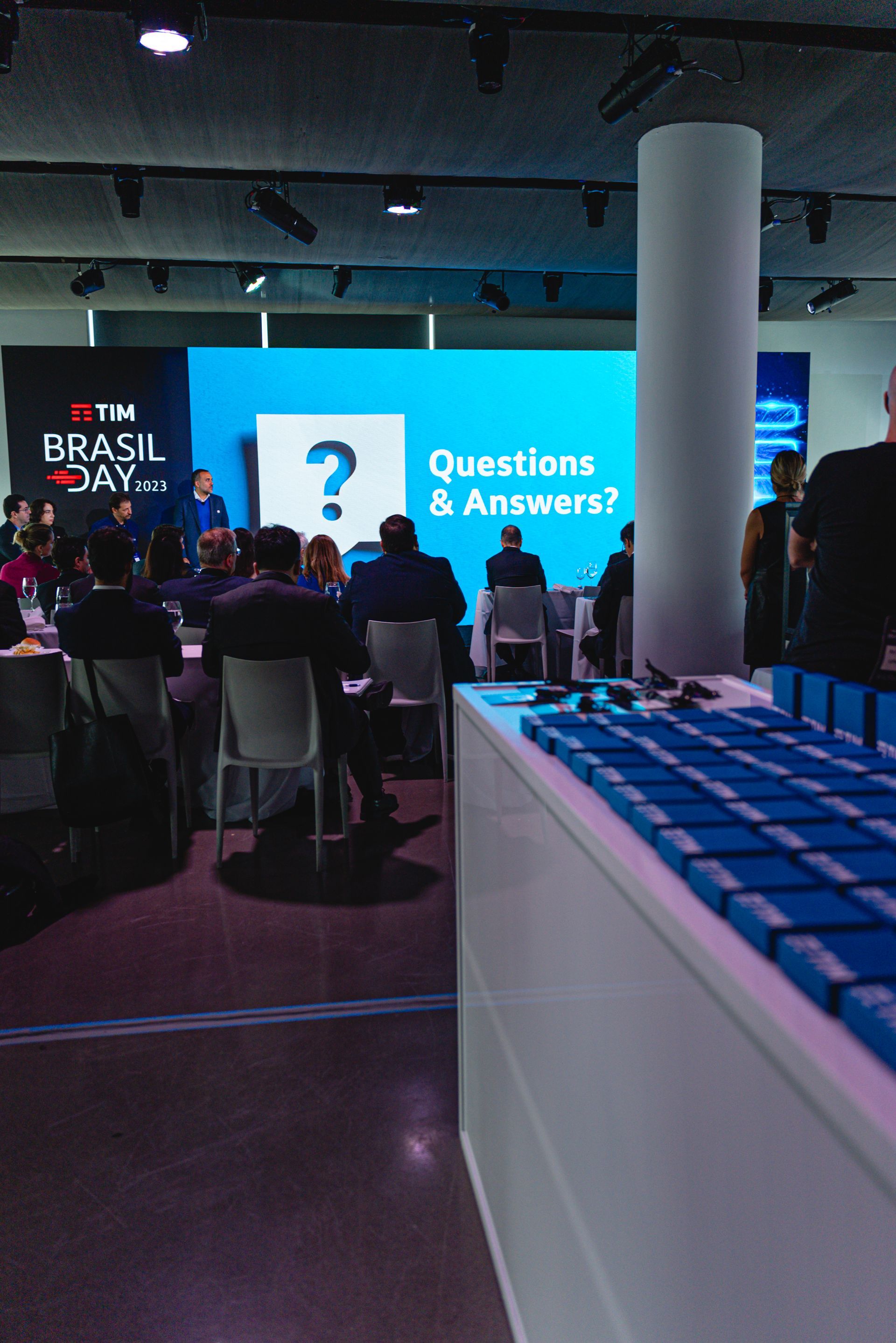 Conference hall with a presentation screen displaying "Questions & Answers?". People seated and standing, some visible logos.