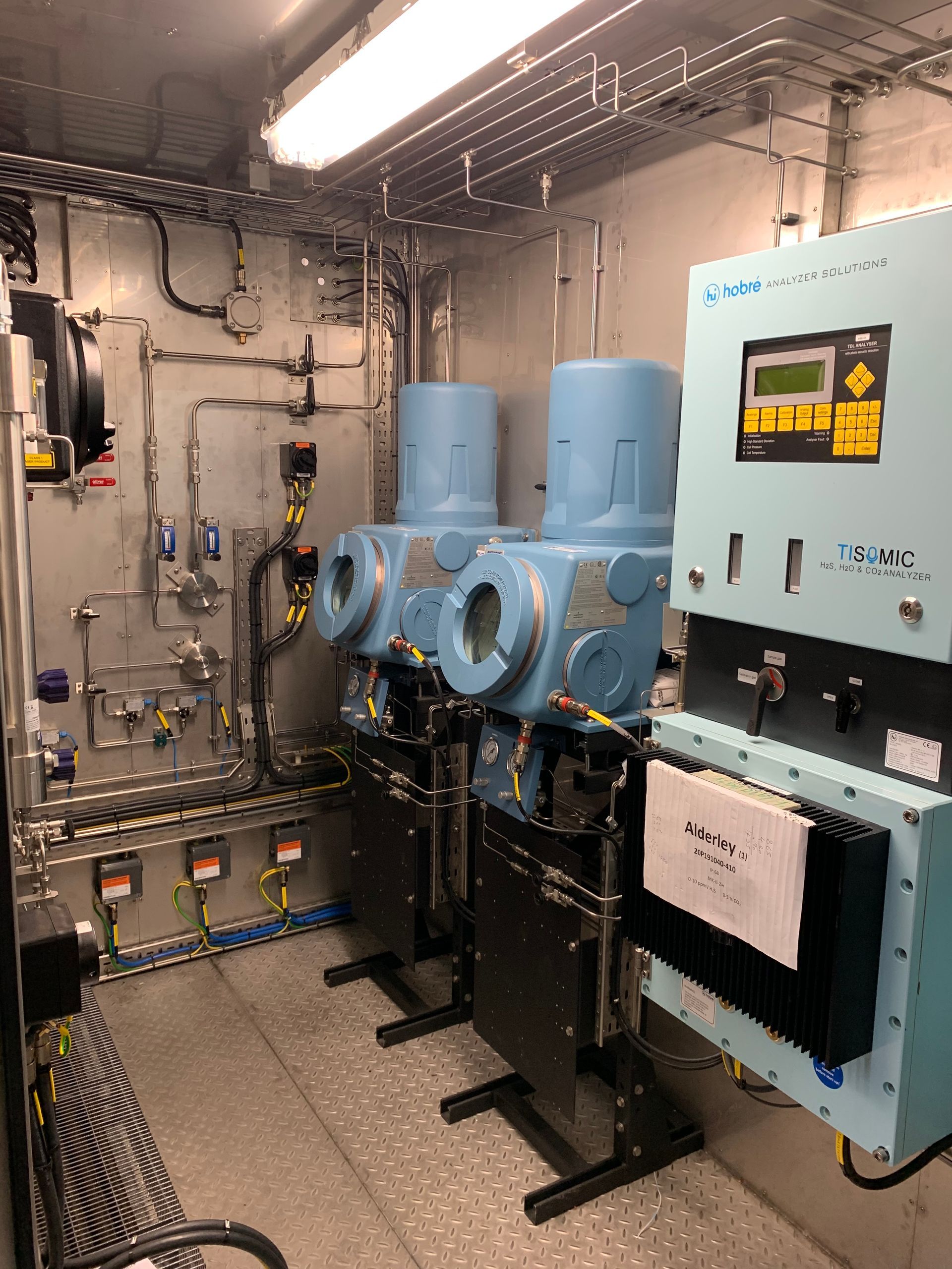 Two light blue industrial sensor housings and a control panel with a digital screen inside a utility room.