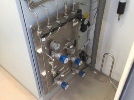 A stainless steel gas manifold panel with pressure gauges, valves, and metal tubing mounted on a wall.