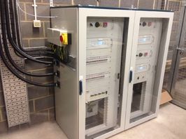 A dual-cabinet industrial electrical distribution panel with glass doors, mounted against a brick wall with cabling.