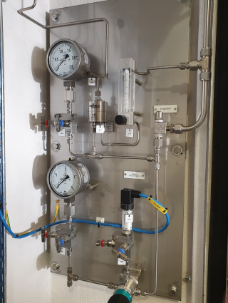 A stainless steel industrial instrument panel mounted on a wall, featuring pressure gauges, valves, and tubing.