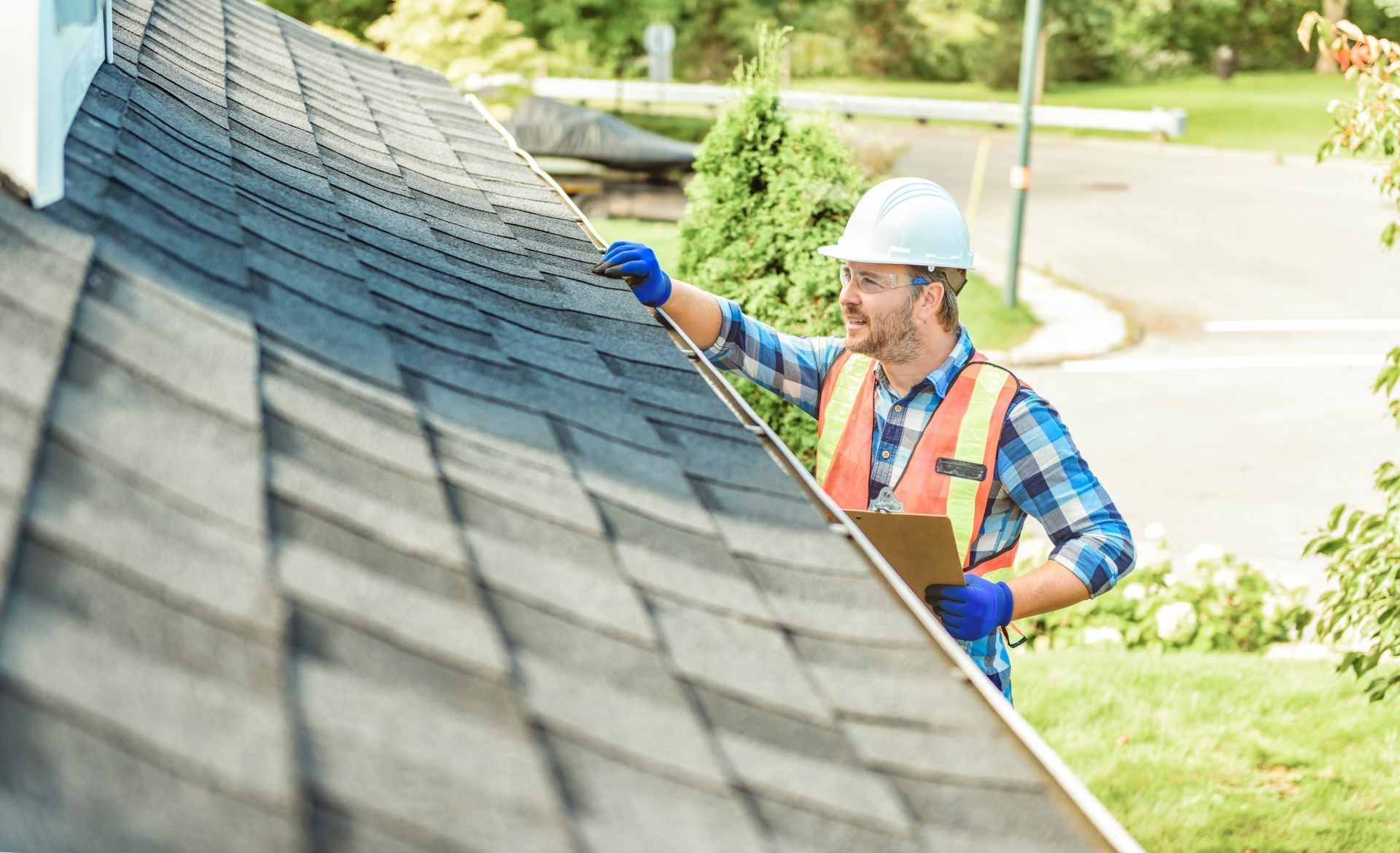 Worker in hard hat and safety vest inspecting a shingled roof with a brush or tool