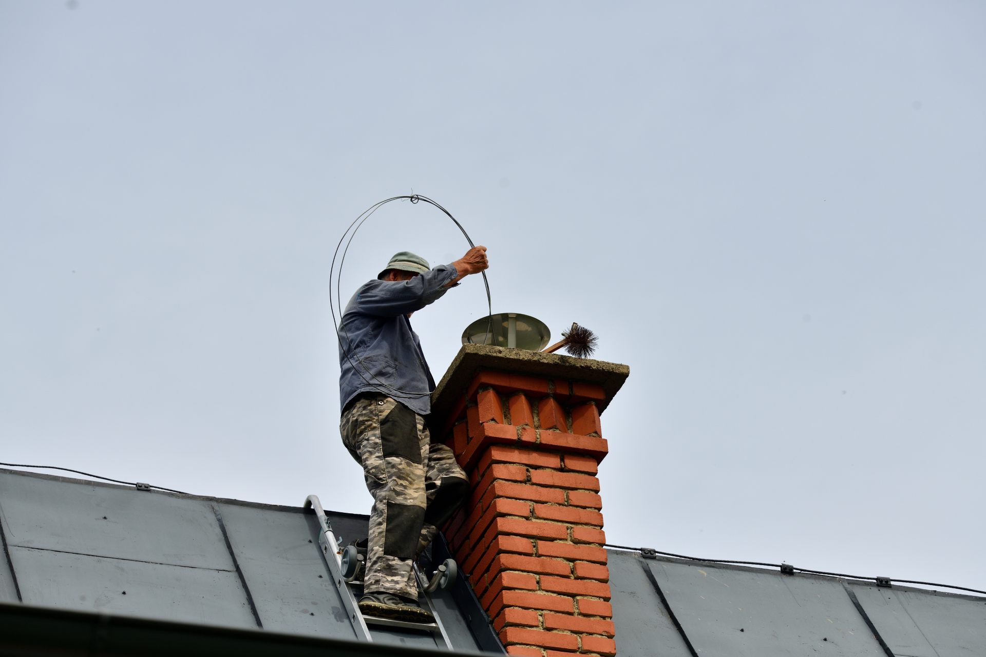 Worker on a roof cleaning a brick chimney with a flexible rod.