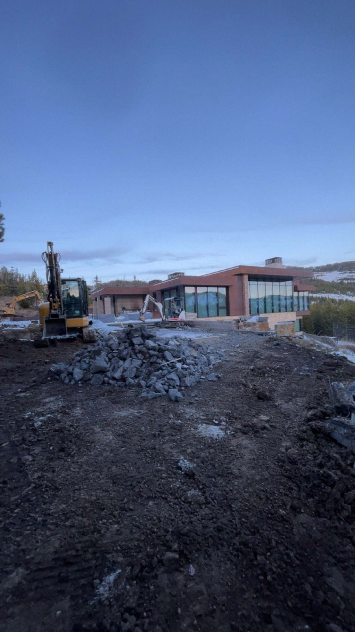 A yellow excavator sits in a muddy, rocky construction site near a modern building with large glass windows at dusk.