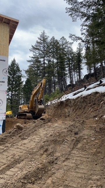 A yellow excavator parked on a dirt hill beside a wood-framed building near a forest.