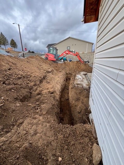 A red excavator works on a deep trench dug into the ground next to a residential house with white siding.