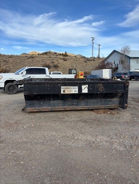 A large, rectangular black metal dumpster sits on a gravel lot in front of a white pickup truck and a building.