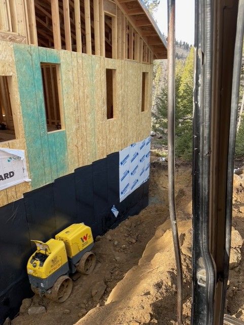 A construction site showing a house frame with partial sheathing and black waterproof foundation coating near a trench.