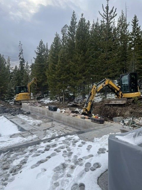 Two yellow excavators work at a snowy construction site near a forest, clearing dirt and rock along a concrete wall.