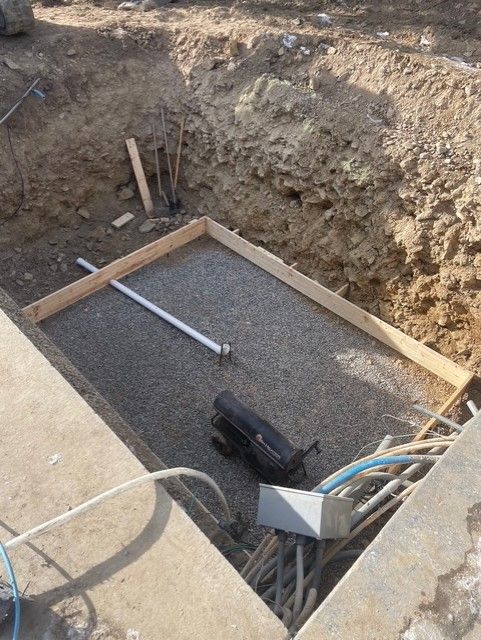 A rectangular trench containing leveled gravel, a wooden frame, a white pipe, and an electrical box with bundled cables.