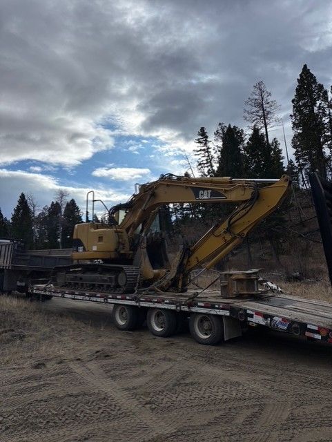 A yellow Caterpillar excavator is loaded onto a flatbed trailer parked on a dirt lot with a treeline in the background.