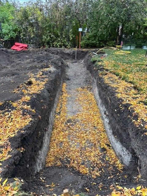 A deep rectangular trench dug into soil in a backyard, partially covered with fallen yellow autumn leaves.