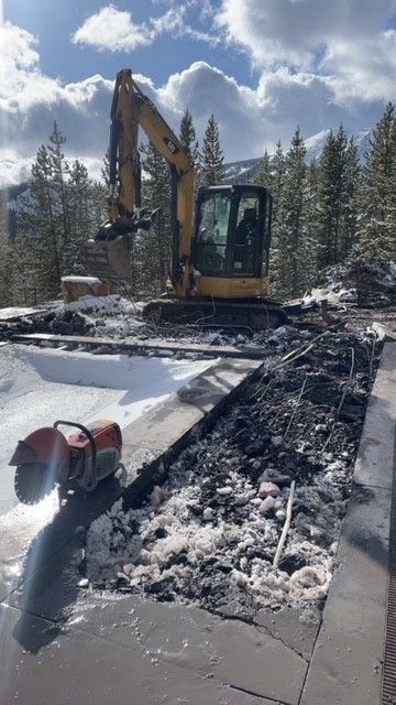 Yellow excavator parked on a snow-covered patio next to a concrete saw during a demolition or construction project.