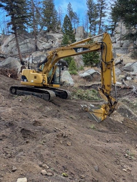 A yellow Caterpillar excavator parked on a dirt slope in a rocky, forested area.