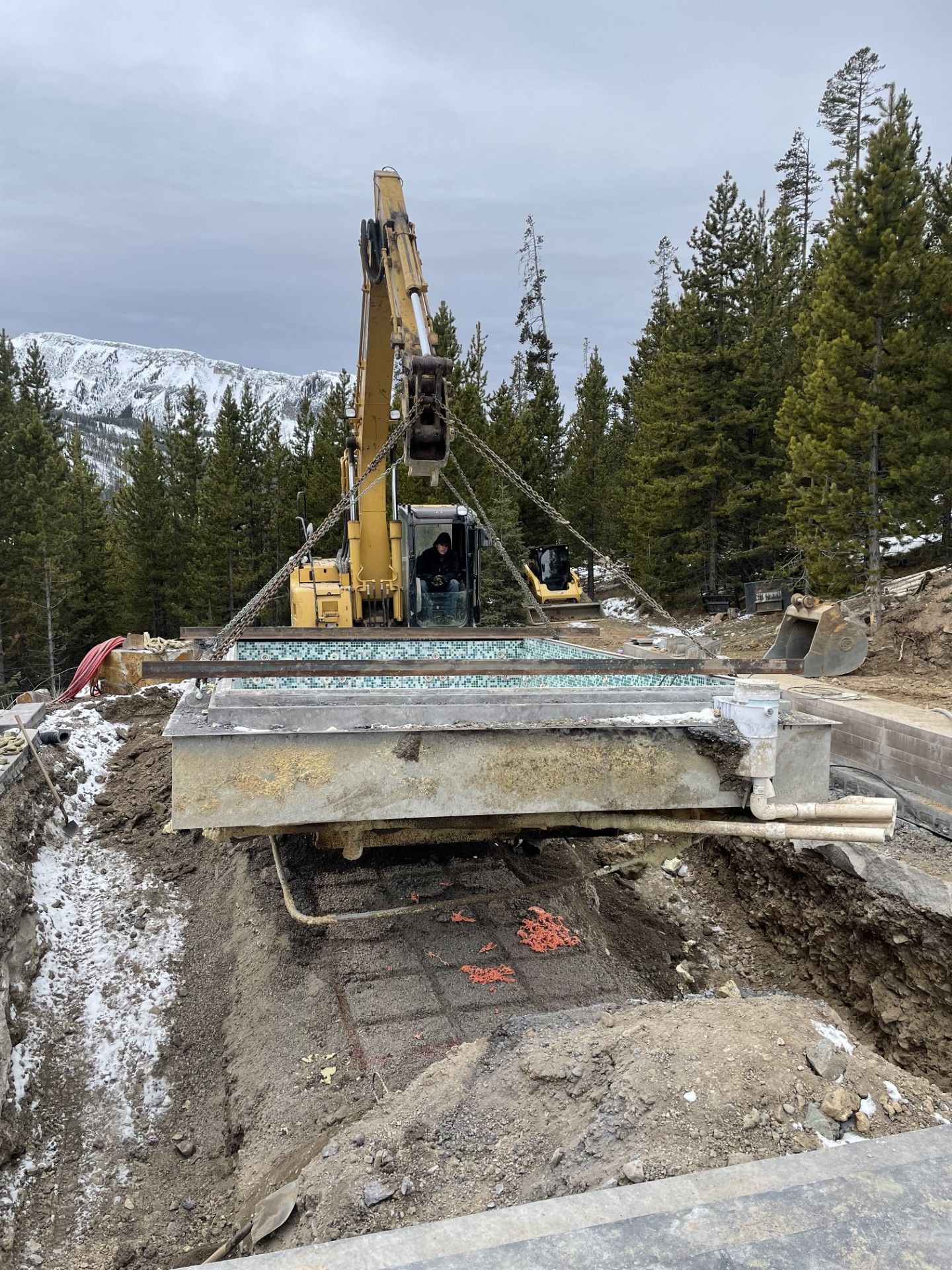 A yellow excavator lowers a precast concrete swimming pool shell into a prepared trench in a snowy, wooded mountain area.