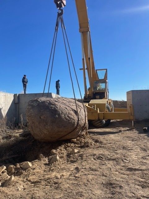A yellow construction crane lifts a large boulder at a rural, sunlit site with two people standing on nearby structures.