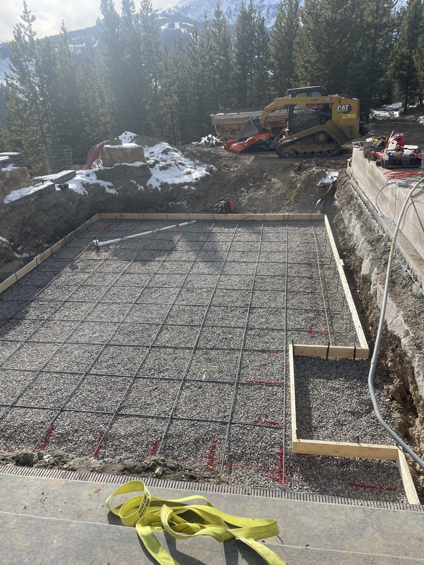 Construction site with a gravel-filled foundation, metal rebar grid, red radiant heating tubes, and a yellow excavator.