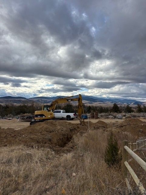 A yellow excavator sits in a dirt lot with a white pickup truck, framed by mountains under a cloudy sky.