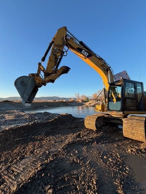 A yellow CAT excavator extends its arm with an empty bucket over a small, calm body of water at a dirt construction site.