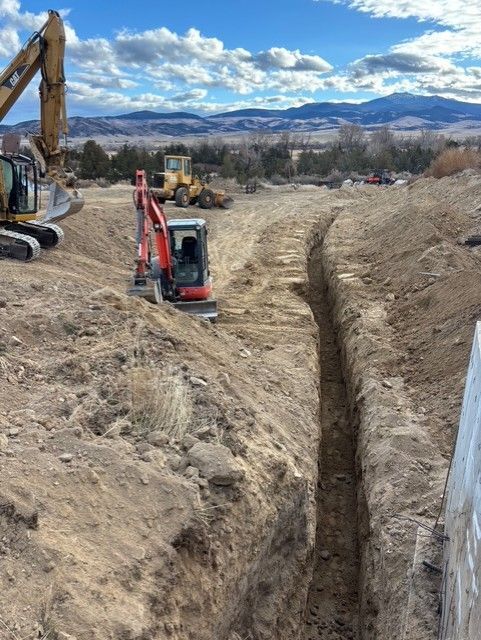 A red excavator sits next to a deep trench on a construction site with mountains in the background.