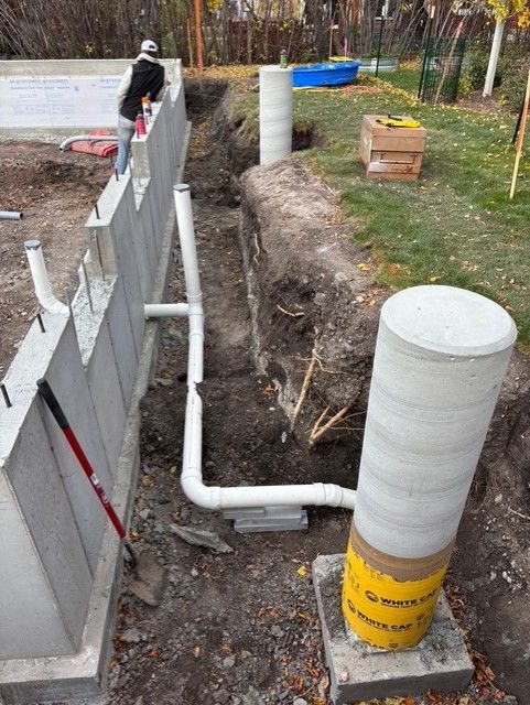 A construction site showing a concrete foundation wall, white PVC drainage pipes, and a vertical concrete pillar.