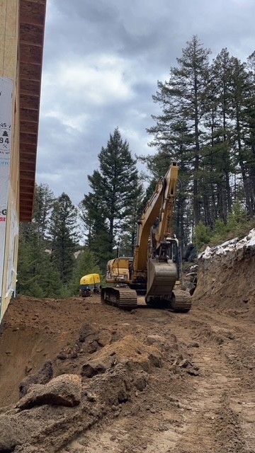 A yellow excavator parked on a dirt construction site next to a building, with pine trees and a cloudy sky in the background.