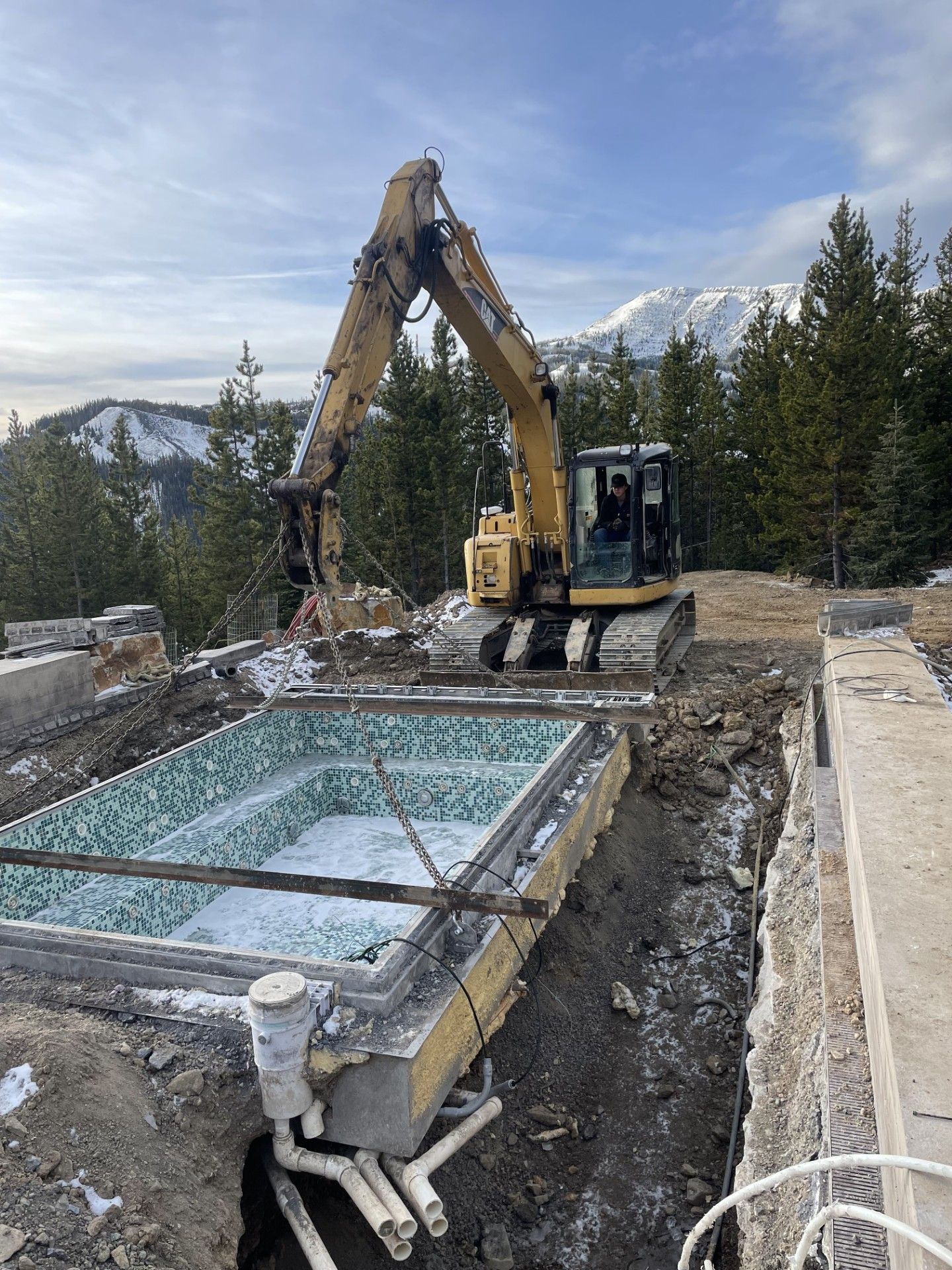 A yellow excavator lifts a tiled, rectangular spa shell from an excavated construction site with mountain views.