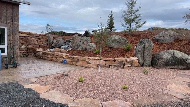 A rustic stone retaining wall borders a gravel garden area next to a wooden building under a cloudy sky.