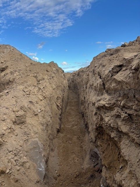 A narrow, deep trench cut into dry, sandy earth under a bright blue sky with scattered clouds.