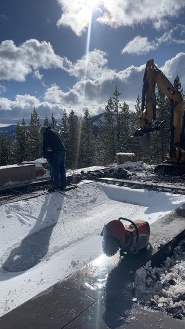 A construction worker and an excavator work on a snow-covered patio overlooking trees and mountains on a sunny day.