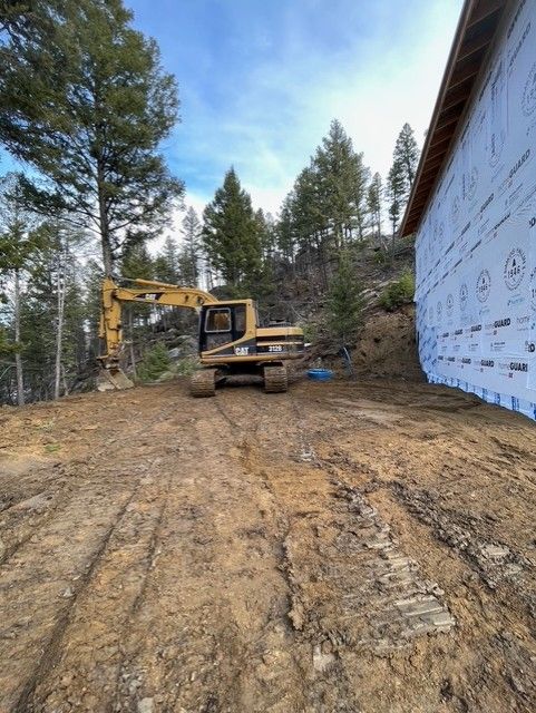 Yellow excavator parked on a dirt lot next to the unfinished, blue-wrapped exterior wall of a new house near a forest.