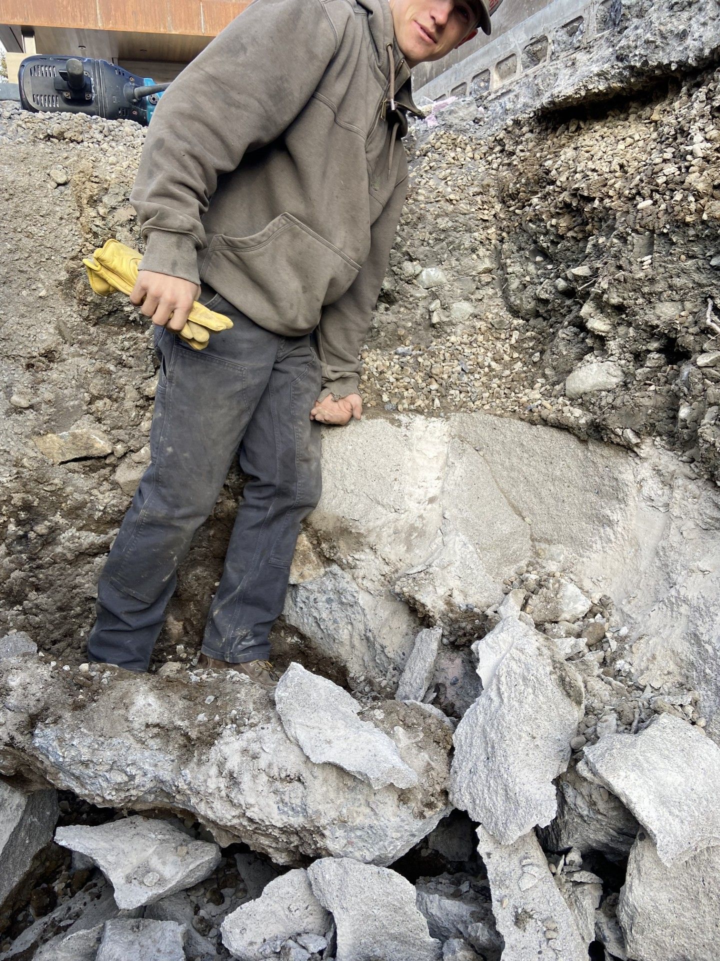 A person in a brown hoodie standing on a pile of concrete rubble at a construction site while holding a yellow tool.