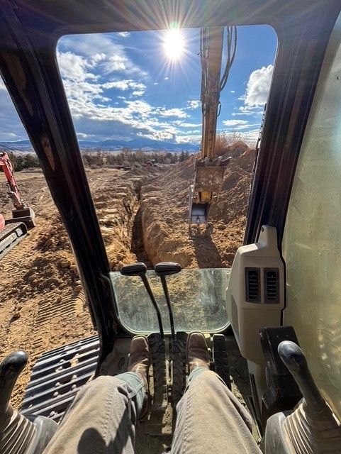 The view from inside an excavator cab looking out over a construction site with an open trench and a bright sunny sky.