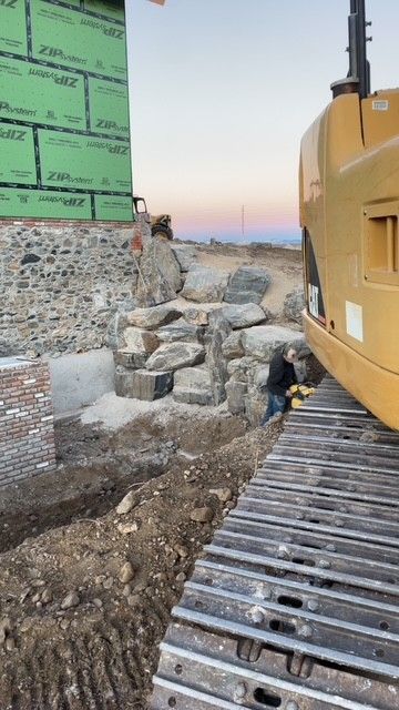 A construction worker stands near a rock wall foundation beside a CAT excavator at a building site under a sunset sky.