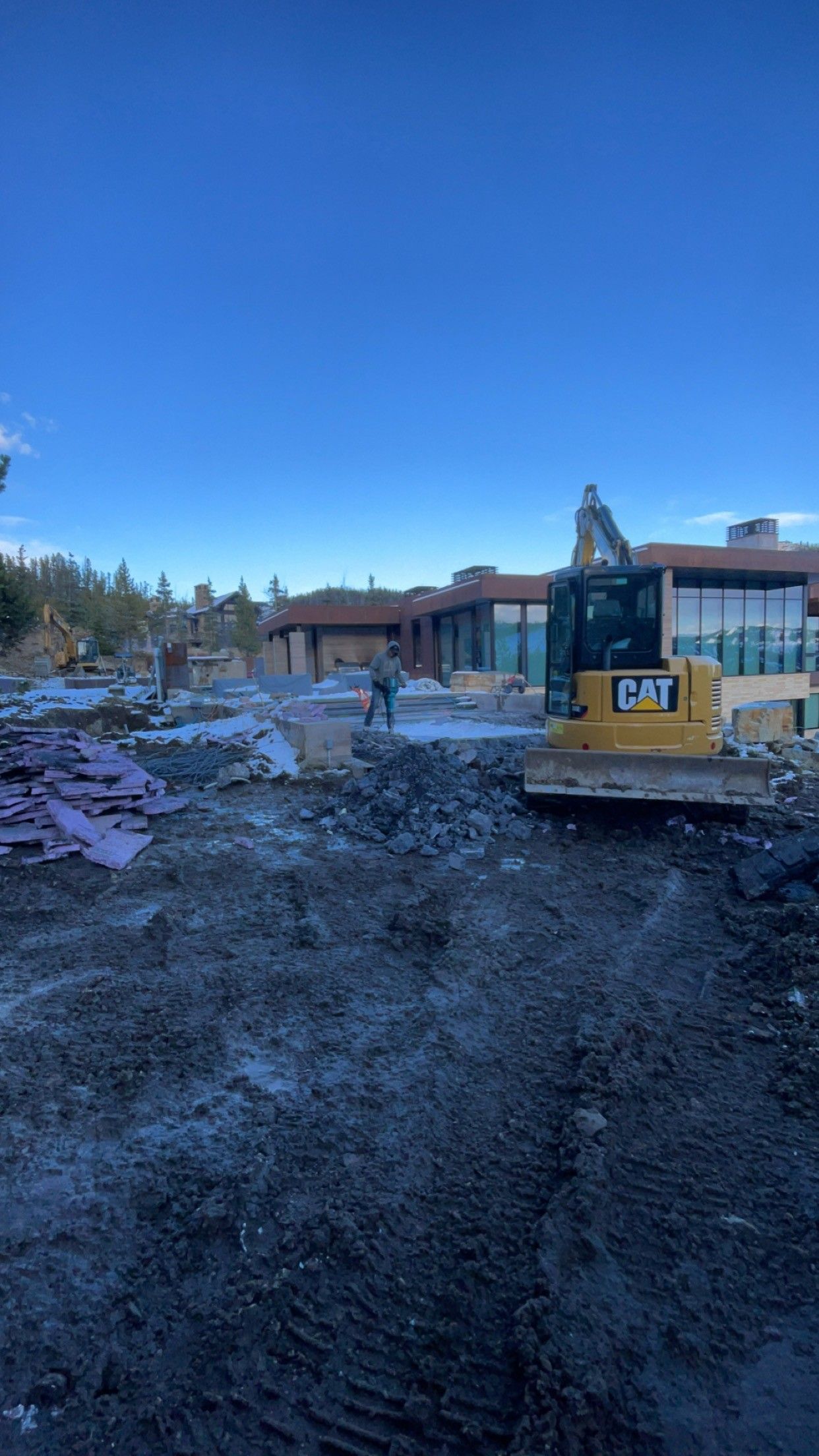 A yellow Caterpillar excavator sits on a dirt construction site in front of a modern house under a clear blue sky.