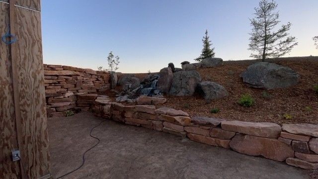 A stone retaining wall surrounds a patio with a rock waterfall feature and landscaping under a clear sky.