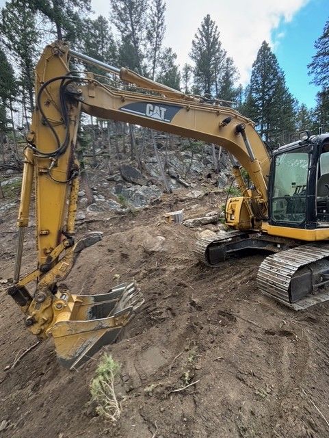 Yellow CAT excavator parked on a sloped, dirt construction site with a forest in the background.