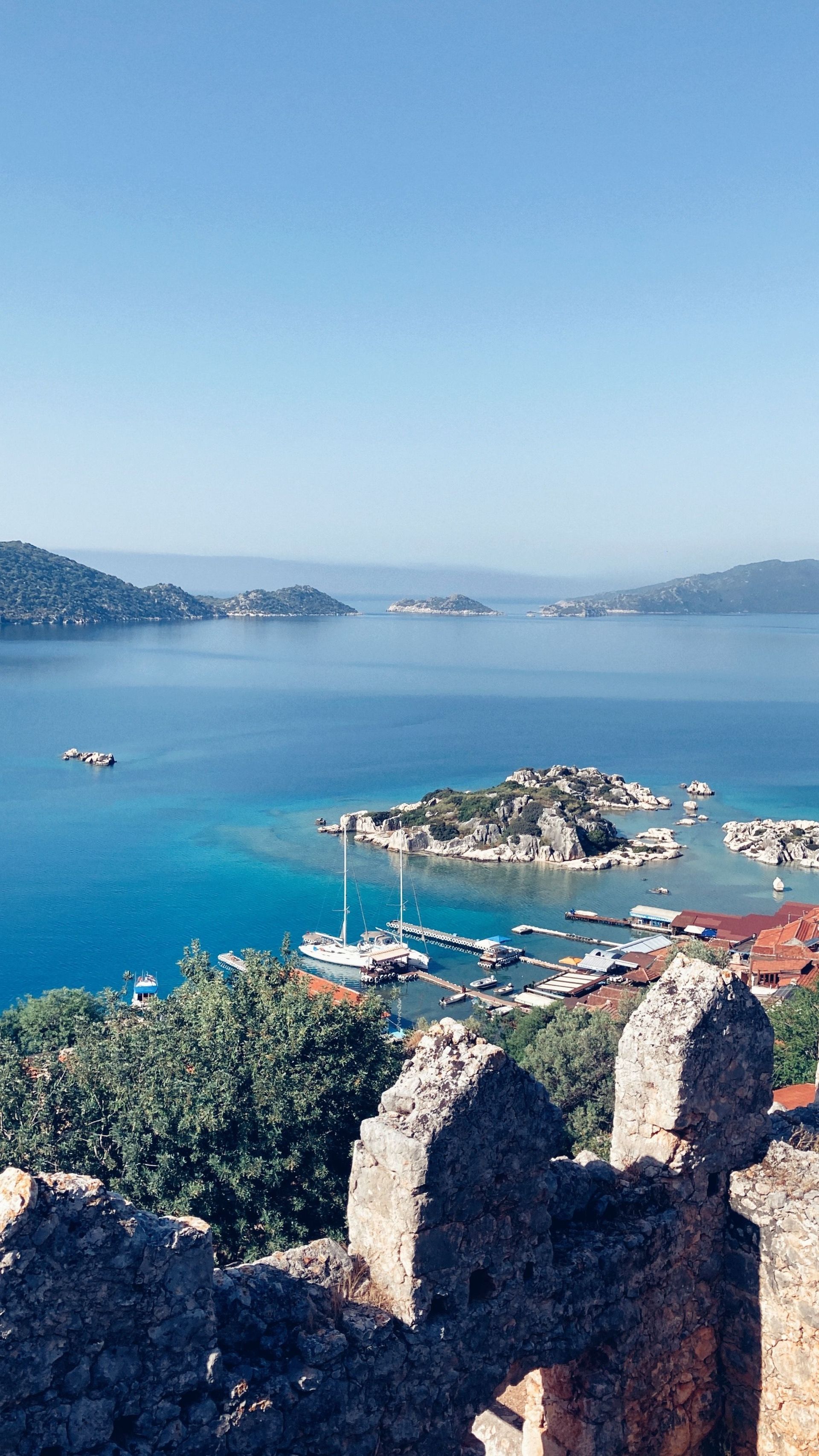 Blue coastal bay with rocky shoreline, small boats, and distant hills under a clear sky