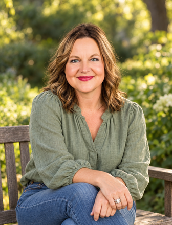 Woman seated on a bench outdoors, smiling in a green blouse and jeans with a blurred garden background