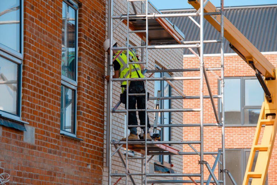A Man is Standing on a Scaffolding Next to a Brick Building — Byron Bay Scaffold in Mullumbimby, NSW