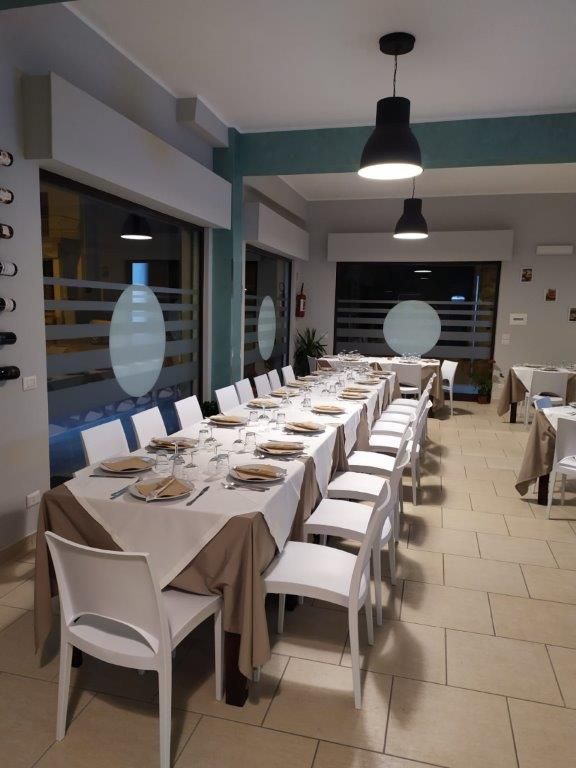 Lungo tavolo da banchetto apparecchiato per una cena in un ristorante moderno, con sedie bianche e lampade a sospensione nere.