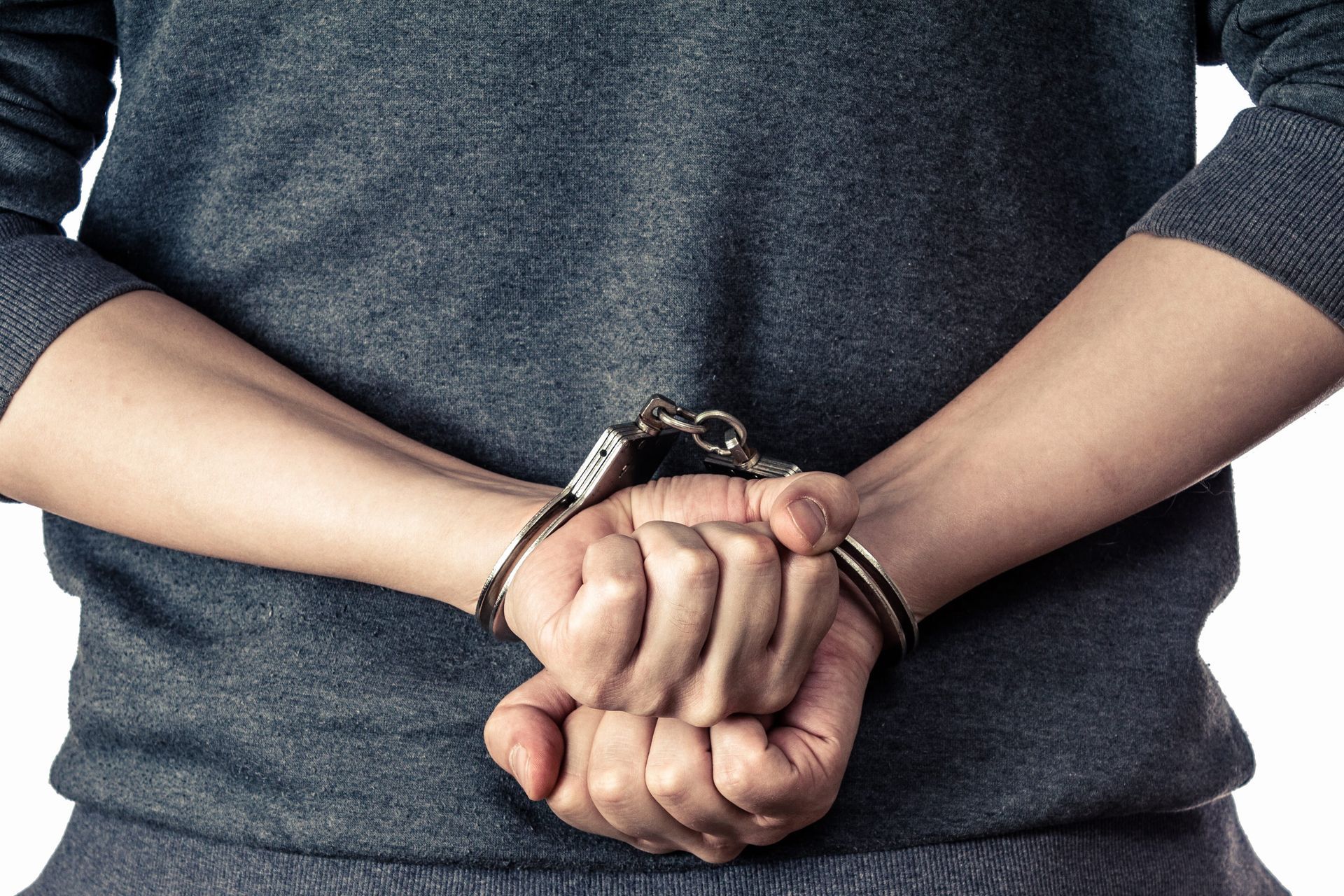 A person in a gray sweater, seen from behind, with their wrists handcuffed behind their back.