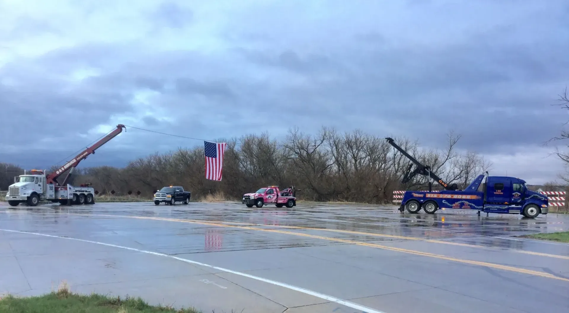 A group of tow trucks are parked on the side of a wet road.