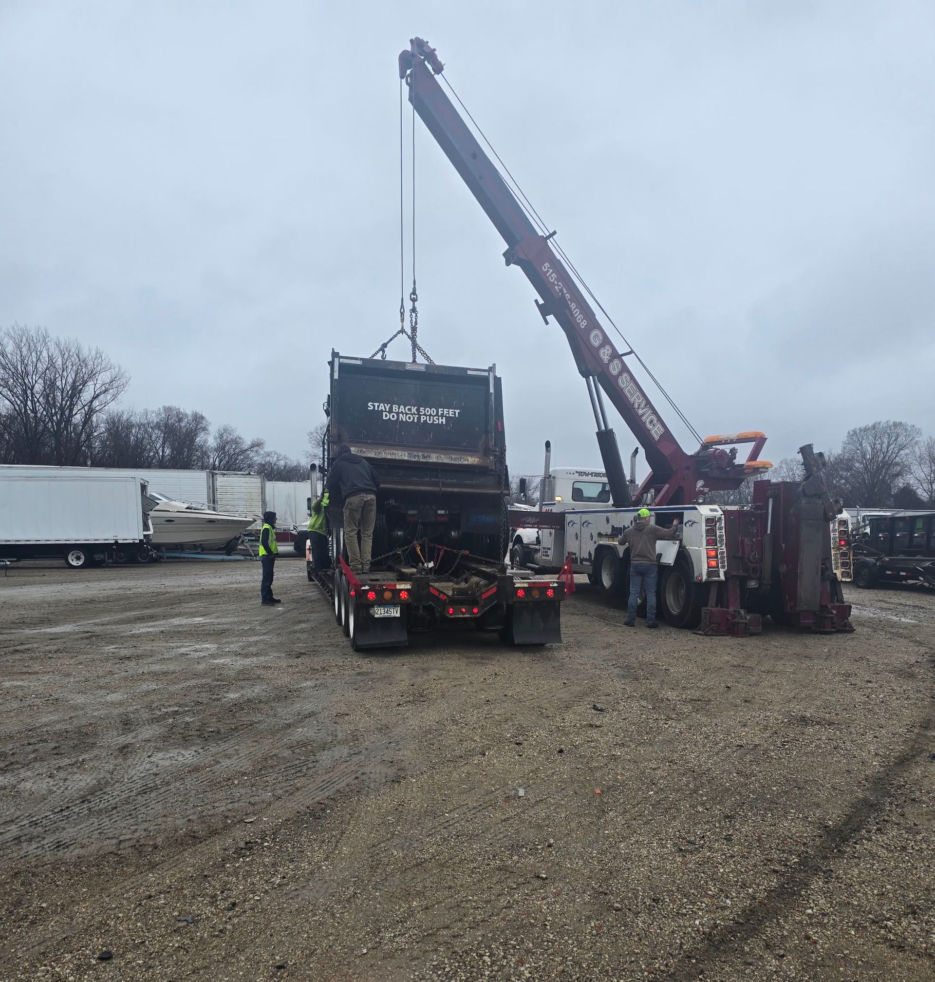 Tow truck lifting a large truck cab in a gravel lot on a cloudy day, with workers nearby