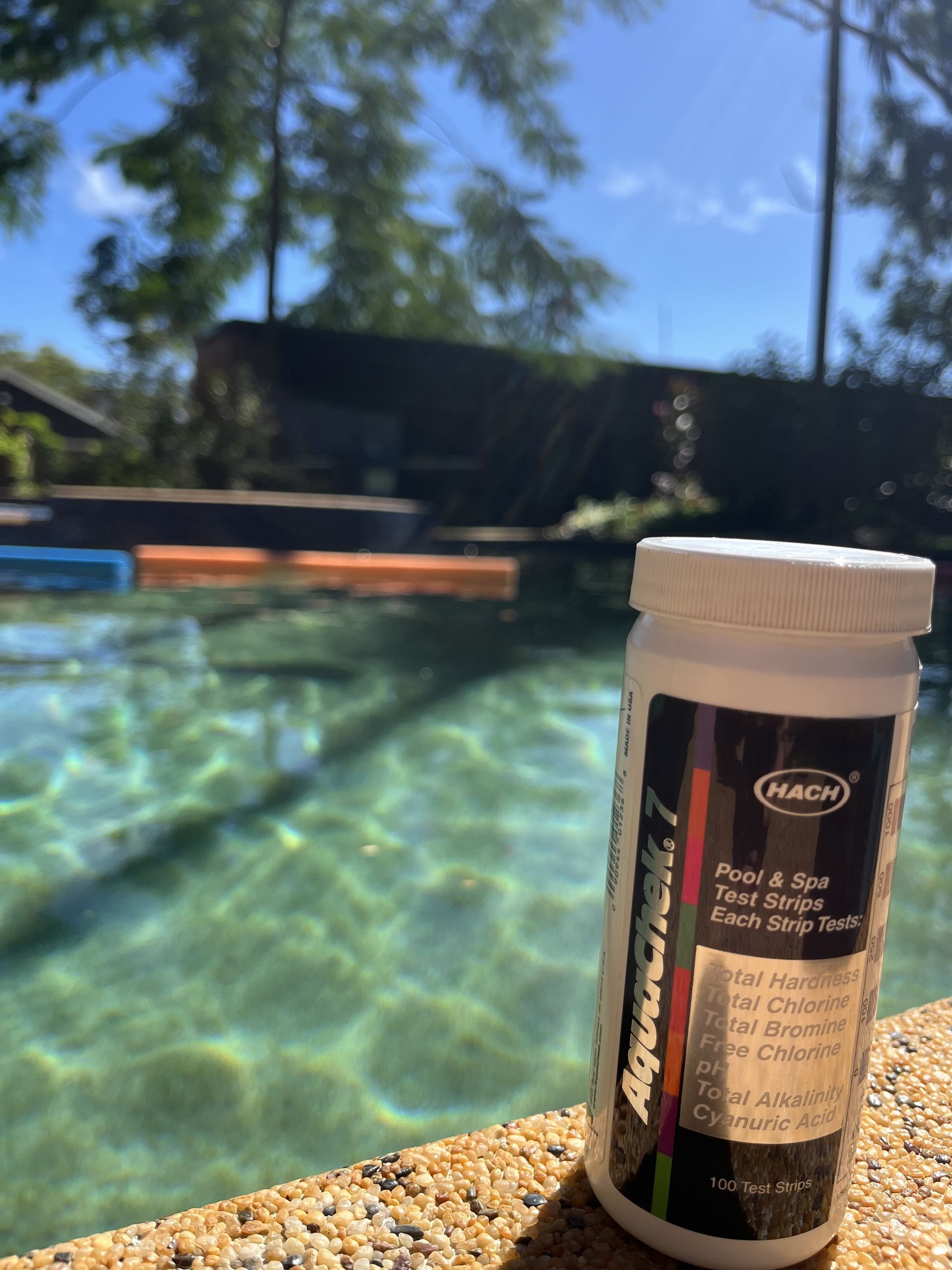 A bottle of Hach AquaChek 7 pool test strips sits on a stone edge beside a sunlit swimming pool — Friendly Pool & Spa Care in Palmerston, NT