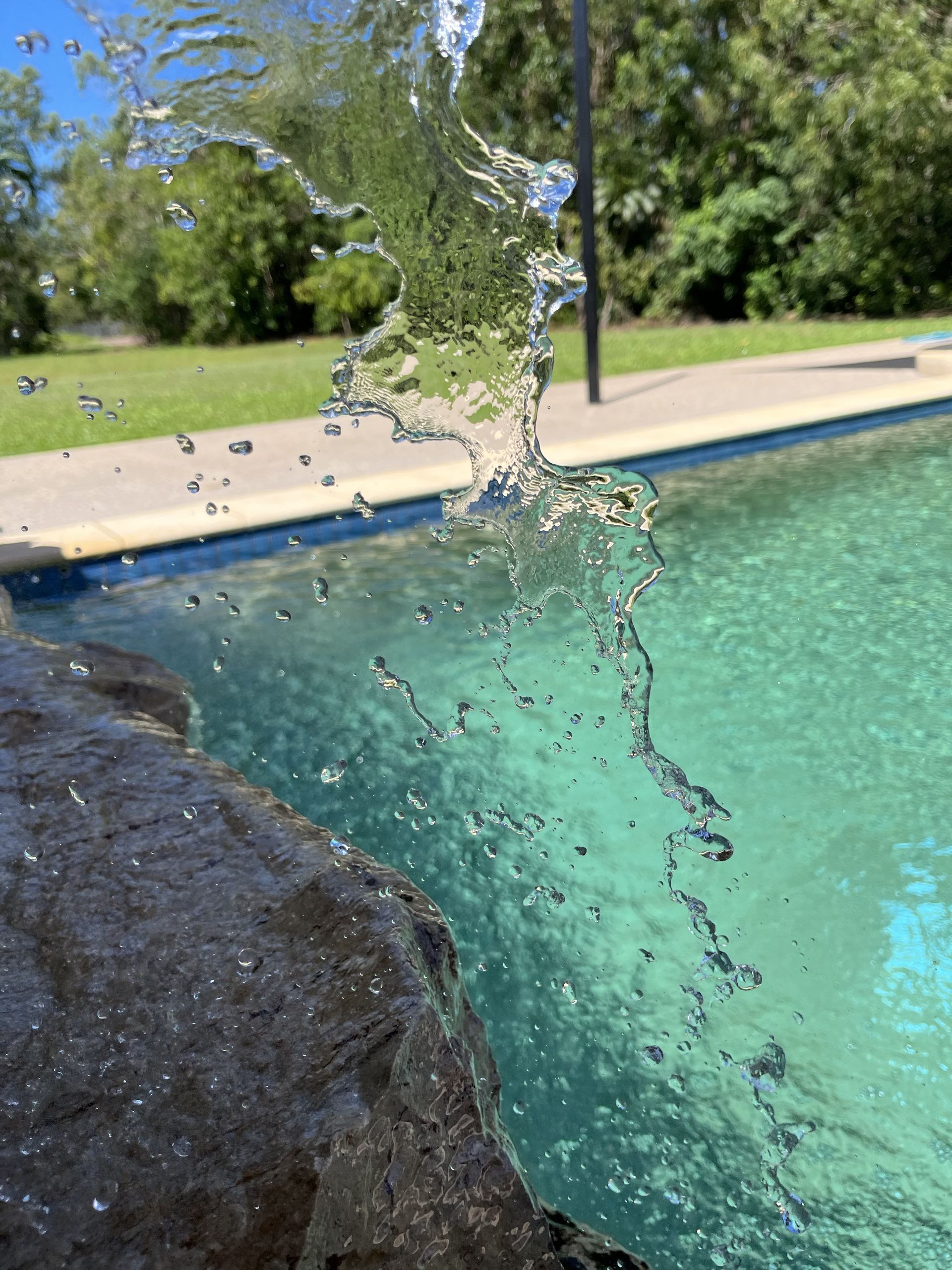 Water cascades from a dark, jagged rock ledge into a turquoise backyard swimming pool under a bright sky — Friendly Pool & Spa Care in Coconut Grove, NT