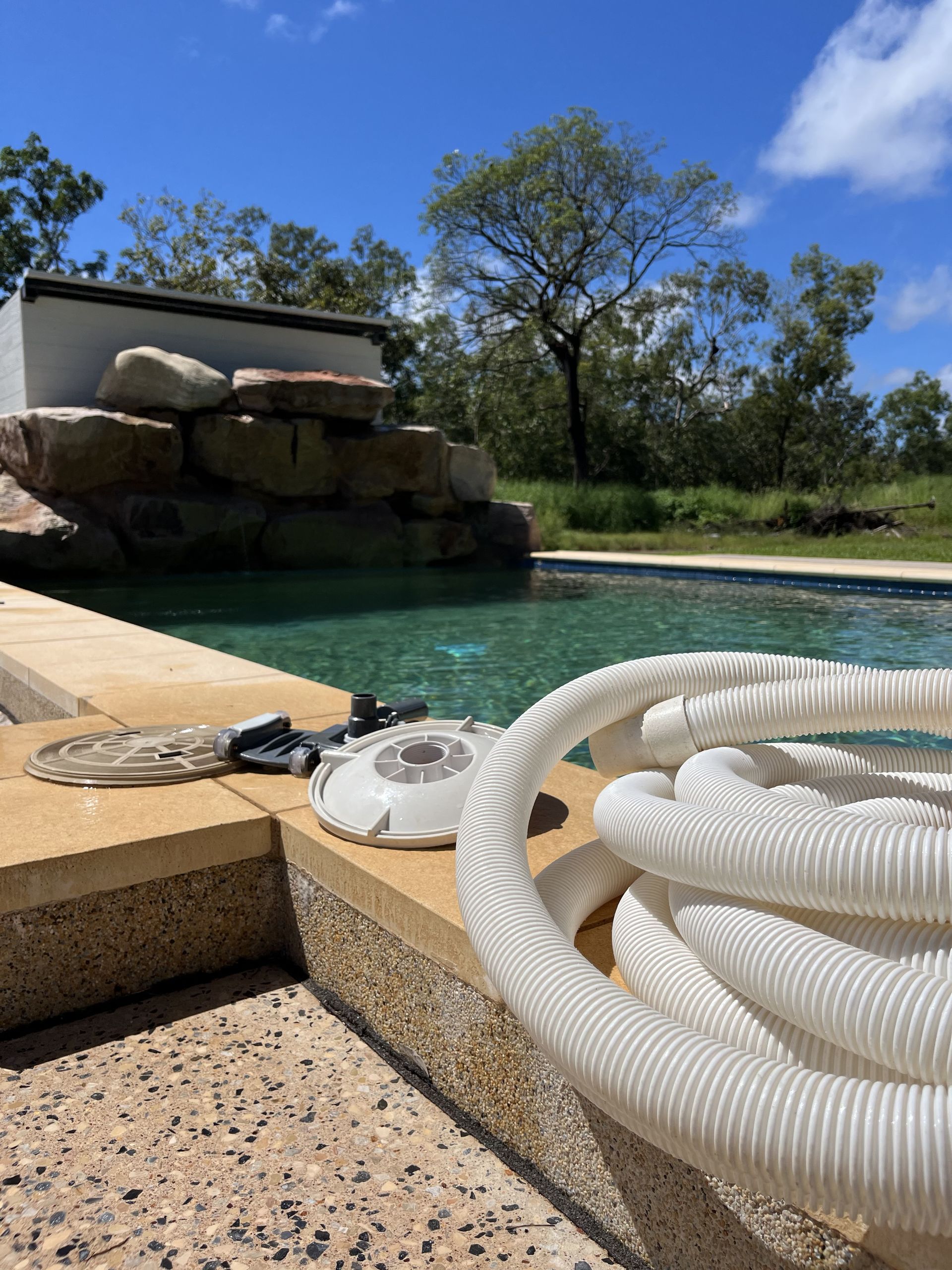 A coiled white pool cleaner hose and equipment lie on a stone poolside next to a rock-lined swimming pool — Friendly Pool & Spa Care in Coconut Grove, NT