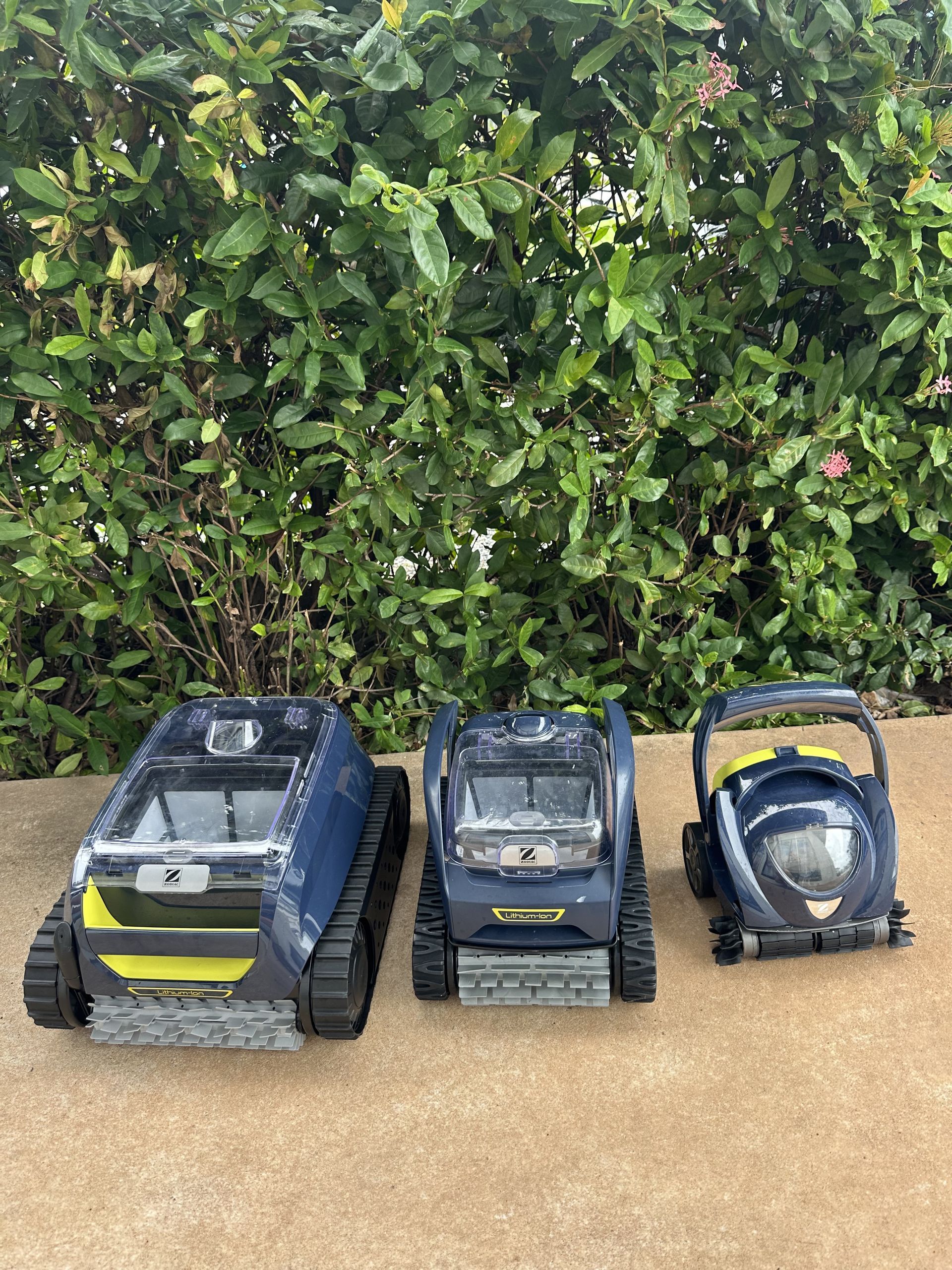 Three pool cleaning robots in varying sizes are lined up on a concrete surface in front of a green hedge — Friendly Pool & Spa Care in Coconut Grove, NT